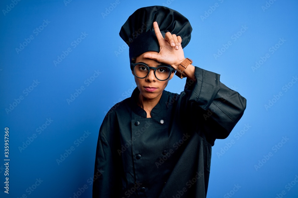 Young african american chef woman wearing cooker uniform and hat over blue background making fun of people with fingers on forehead doing loser gesture mocking and insulting. Young african american chef woman wearing cooker uniform and hat over blue background making fun of people with fingers on forehead doing loser gesture mocking and insulting.