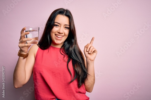 Young brunette woman drinking a glass of fresh water over pink isolated background surprised with an idea or question pointing finger with happy face, number one