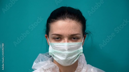 Beautiful Caucasian pretty girl with black hair in white protective suit on blue background puts respirator, glasses and two medical masks from virus on her face, zips jacket, put on a hood close up.  photo