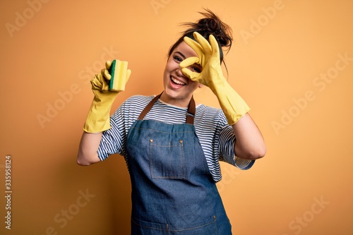 Young brunette cleaner woman wearing housekeeping gloves holding scourer scrub with happy face smiling doing ok sign with hand on eye looking through fingers photo