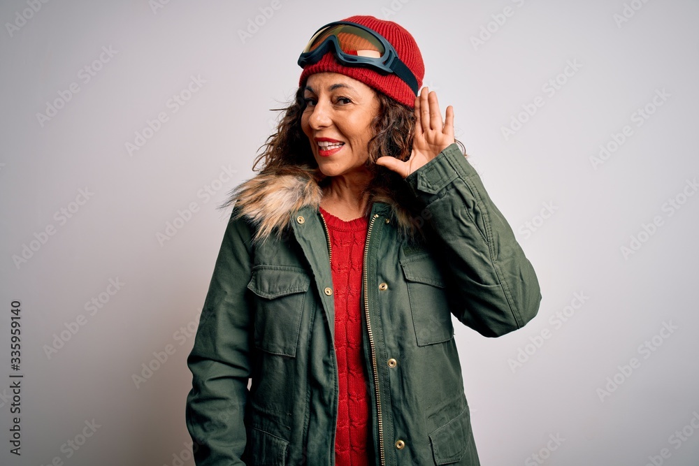 Middle age skier woman wearing snow sportswear and ski goggles over white background smiling with hand over ear listening an hearing to rumor or gossip. Deafness concept. Middle age skier woman wearing snow sportswear and ski goggles over white background smiling with hand over ear listening an hearing to rumor or gossip. Deafness concept.