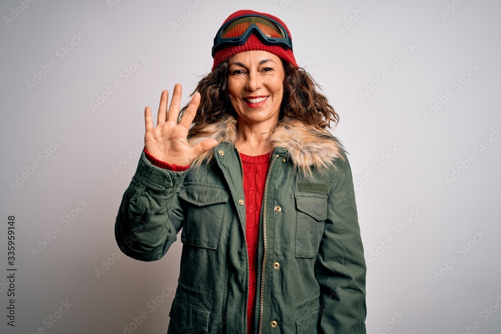 Middle age skier woman wearing snow sportswear and ski goggles over white background showing and pointing up with fingers number five while smiling confident and happy. Middle age skier woman wearing snow sportswear and ski goggles over white background showing and pointing up with fingers number five while smiling confident and happy.
