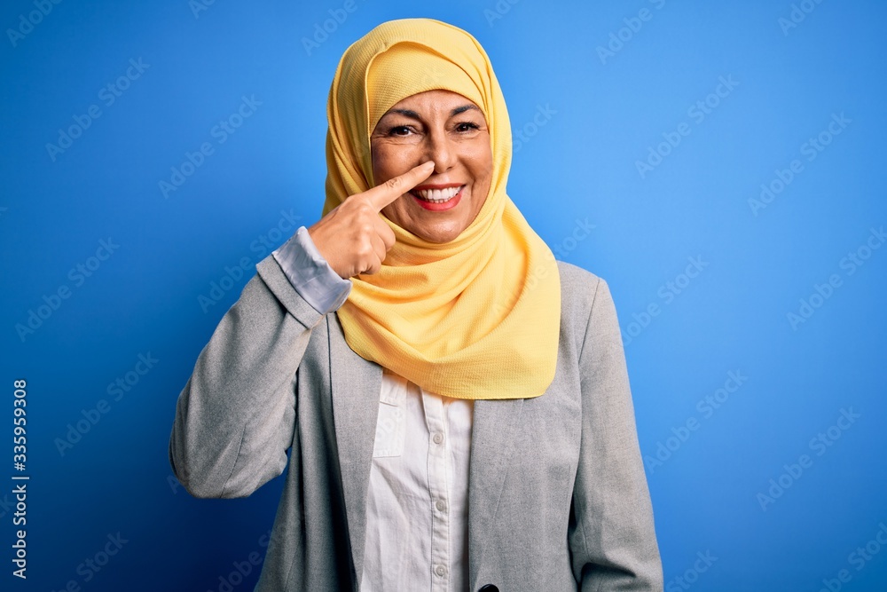 Middle age brunette business woman wearing muslim traditional hijab over blue background Pointing with hand finger to face and nose, smiling cheerful. Beauty concept Middle age brunette business woman wearing muslim traditional hijab over blue background Pointing with hand finger to face and nose, smiling cheerful. Beauty concept