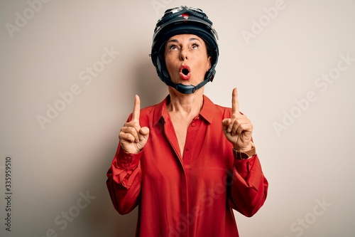 Middle age motorcyclist woman wearing motorcycle helmet over isolated white background amazed and surprised looking up and pointing with fingers and raised arms.