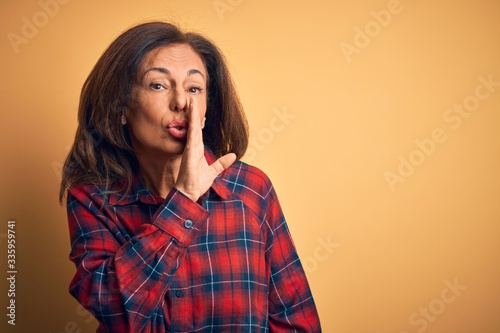 Middle age beautiful woman wearing casual shirt standing over isolated yellow background hand on mouth telling secret rumor, whispering malicious talk conversation