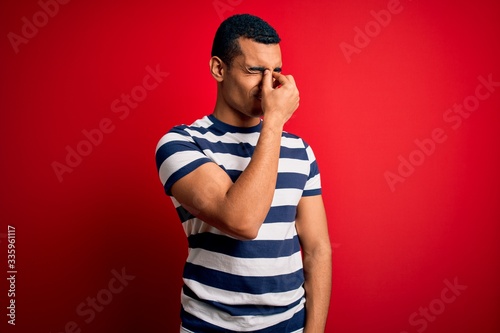 Handsome african american man wearing casual striped t-shirt standing over red background tired rubbing nose and eyes feeling fatigue and headache. Stress and frustration concept.