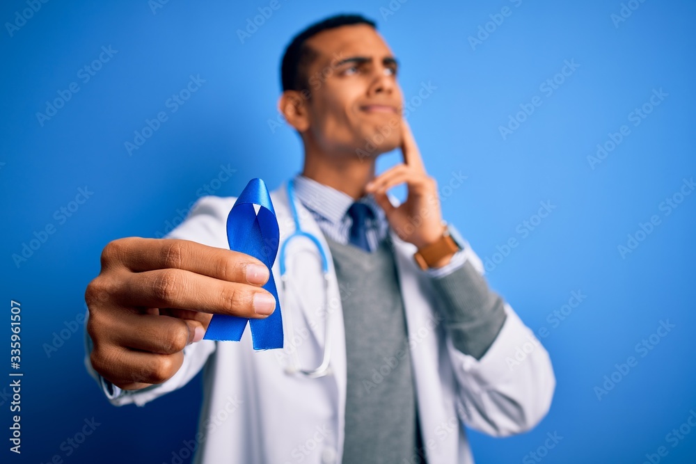 Young handsome african american doctor man holding blue cancer ribbon symbol serious face thinking about question, very confused idea