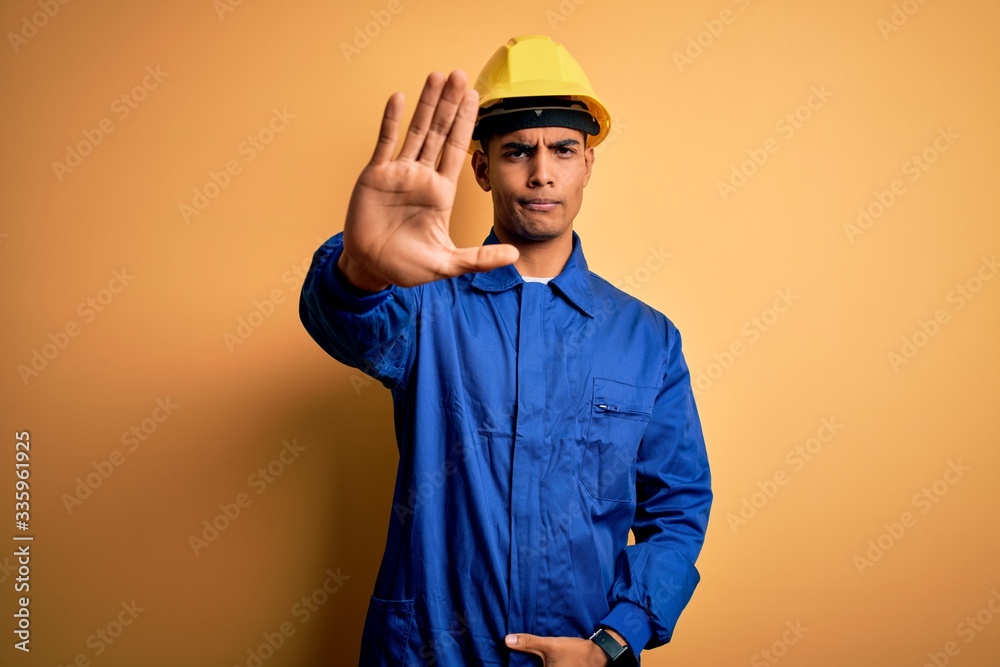 Young handsome african american worker man wearing blue uniform and security helmet doing stop sing with palm of the hand. Warning expression with negative and serious gesture on the face.