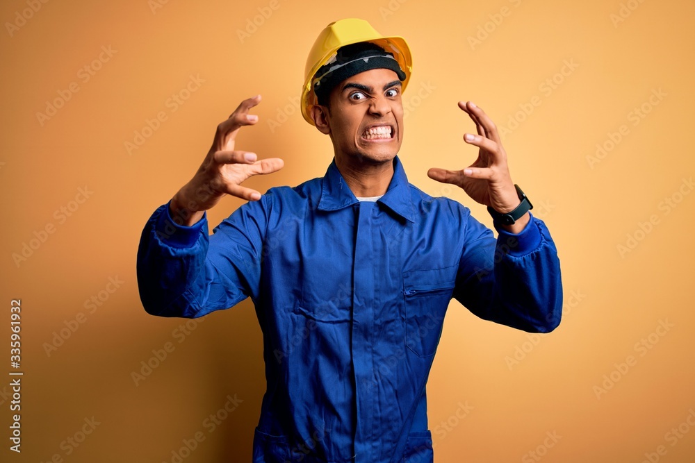 Young handsome african american worker man wearing blue uniform and security helmet Shouting frustrated with rage, hands trying to strangle, yelling mad Young handsome african american worker man wearing blue uniform and security helmet Shouting frustrated with rage, hands trying to strangle, yelling mad