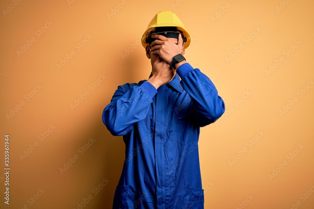 Young handsome african american worker man wearing blue uniform and security helmet Covering eyes and mouth with hands, surprised and shocked. Hiding emotion