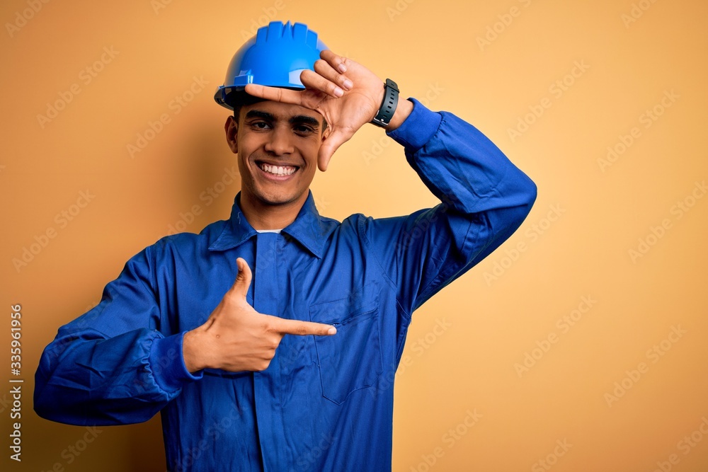 Young handsome african american worker man wearing blue uniform and security helmet smiling making frame with hands and fingers with happy face. Creativity and photography concept.
