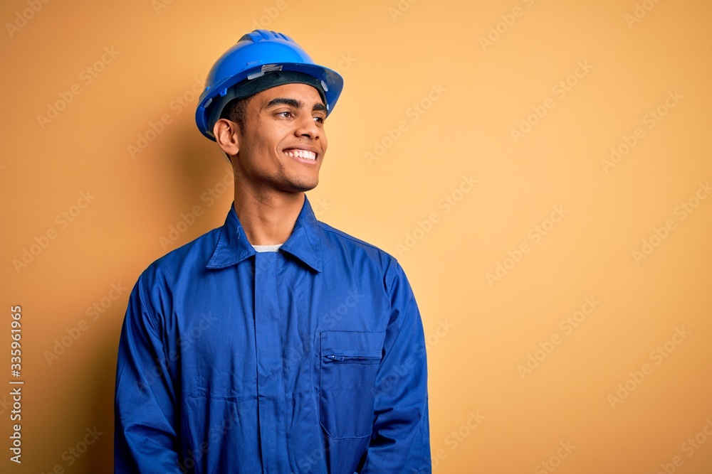 Young handsome african american worker man wearing blue uniform and security helmet looking away to side with smile on face, natural expression. Laughing confident.