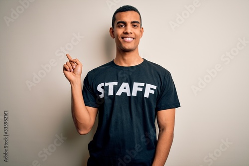 Young handsome african american worker man wearing staff uniform over white background cheerful with a smile on face pointing with hand and finger up to the side with happy and natural expression © Krakenimages.com