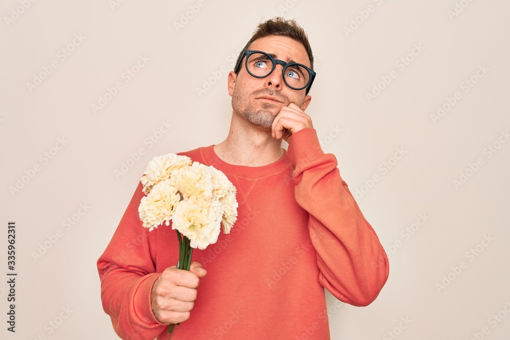 Young handsome man with blue eyes holding bouquet of flowers over white background serious face thinking about question, very confused idea Young handsome man with blue eyes holding bouquet of flowers over white background serious face thinking about question, very confused idea
