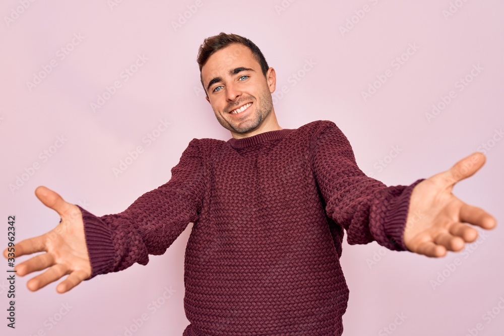 Young handsome man with blue eyes wearing casual sweater standing over pink background smiling cheerful offering hands giving assistance and acceptance. Young handsome man with blue eyes wearing casual sweater standing over pink background smiling cheerful offering hands giving assistance and acceptance.