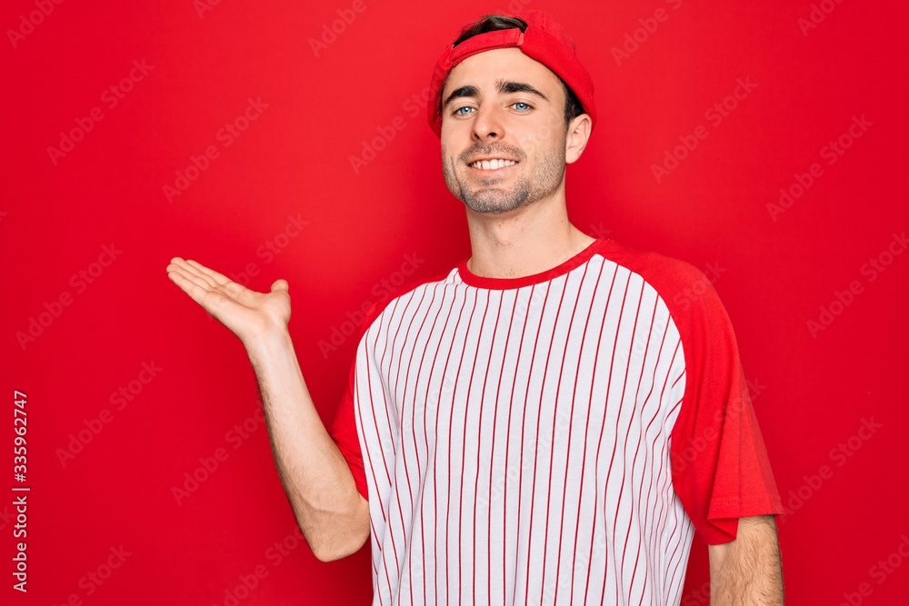 Young handsome sporty man with blue eyes wearing striped baseball t-shirt and cap smiling cheerful presenting and pointing with palm of hand looking at the camera.