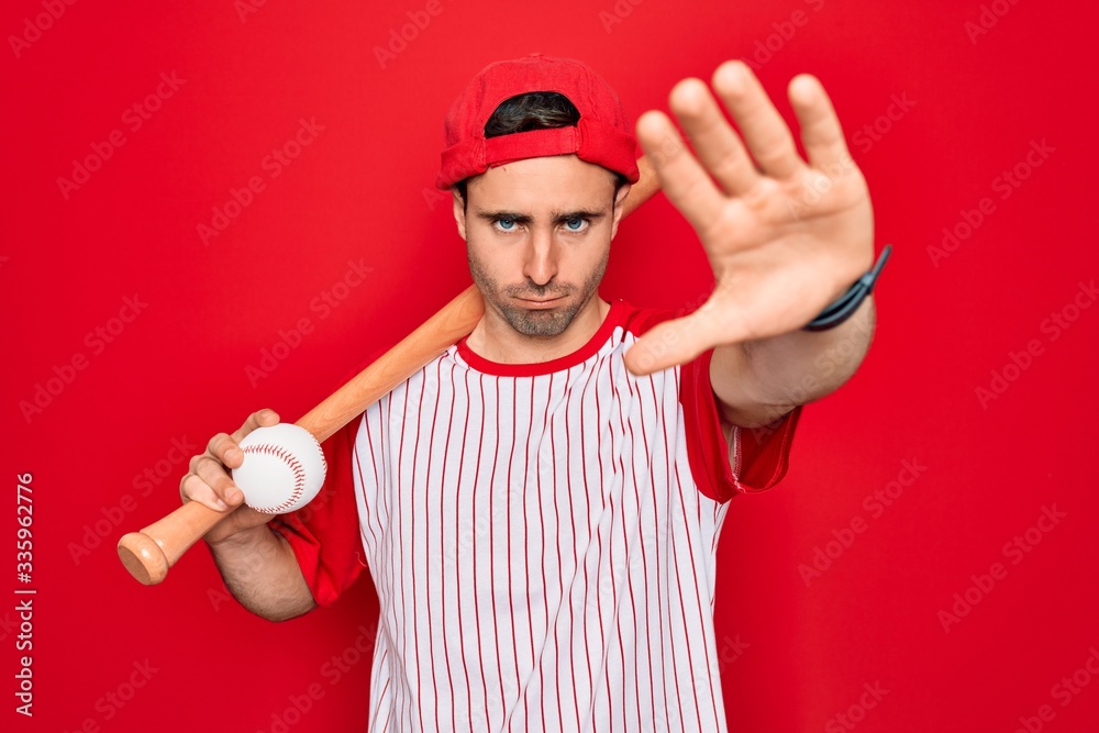 Young handsome sporty man with blue eyes playing baseball wearing cap holding bat and ball with open hand doing stop sign with serious and confident expression, defense gesture Young handsome sporty man with blue eyes playing baseball wearing cap holding bat and ball with open hand doing stop sign with serious and confident expression, defense gesture
