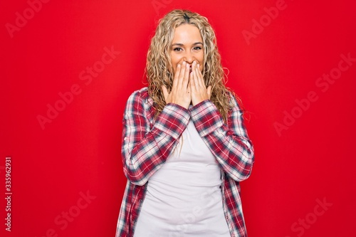 Young beautiful blonde woman wearing casual shirt standing over isolated red background laughing and embarrassed giggle covering mouth with hands, gossip and scandal concept