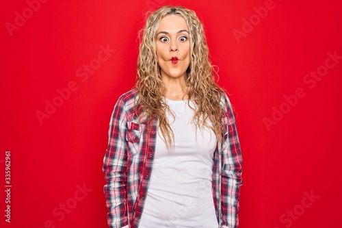 Young beautiful blonde woman wearing casual shirt standing over isolated red background making fish face with lips, crazy and comical gesture. Funny expression.