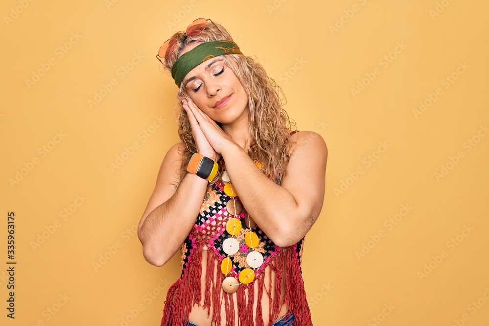 Beautiful blonde hippie woman wearing sunglasses and accessories over yellow background sleeping tired dreaming and posing with hands together while smiling with closed eyes.