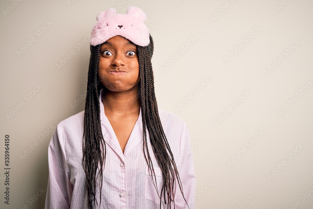 Young african american woman wearing pink pajama and sleep mask over isolated background puffing cheeks with funny face. Mouth inflated with air, crazy expression. Young african american woman wearing pink pajama and sleep mask over isolated background puffing cheeks with funny face. Mouth inflated with air, crazy expression.