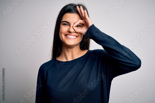 Young beautiful brunette woman wearing casual sweater standing over white background doing ok gesture with hand smiling, eye looking through fingers with happy face.