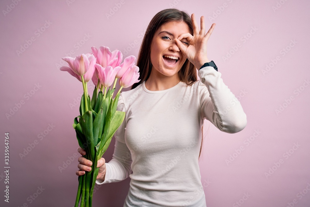 Young beautiful romantic woman with blue eyes holding beauty bouquet of pink tulips with happy face smiling doing ok sign with hand on eye looking through fingers Young beautiful romantic woman with blue eyes holding beauty bouquet of pink tulips with happy face smiling doing ok sign with hand on eye looking through fingers