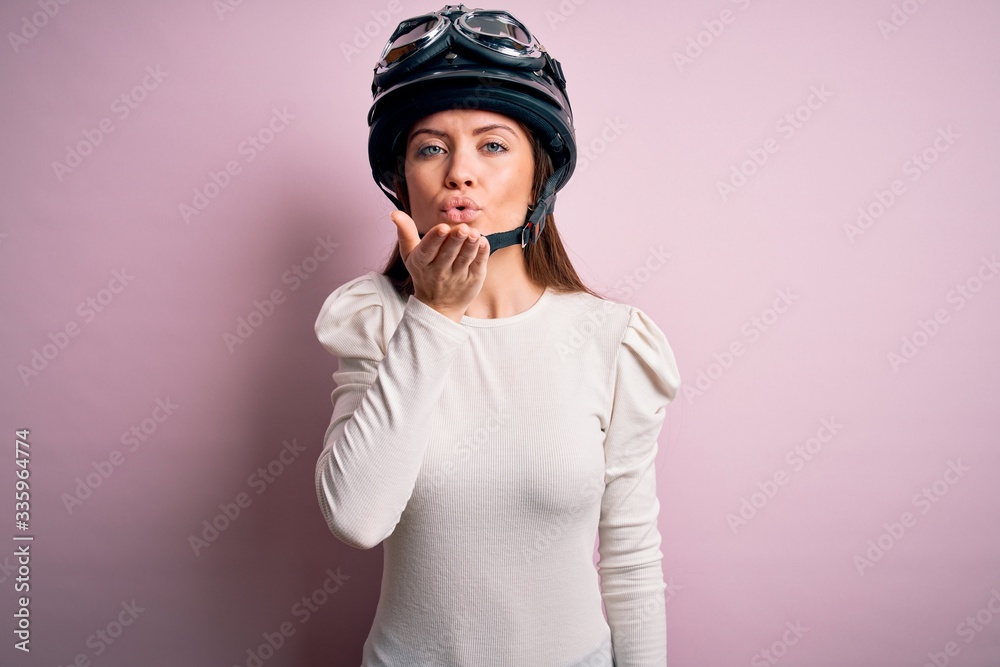 Young beautiful motorcyclist woman with blue eyes wearing moto helmet over pink background looking at the camera blowing a kiss with hand on air being lovely and sexy. Love expression. Young beautiful motorcyclist woman with blue eyes wearing moto helmet over pink background looking at the camera blowing a kiss with hand on air being lovely and sexy. Love expression.