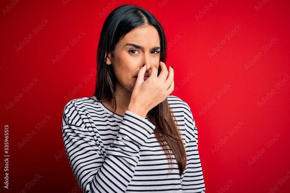 Young beautiful brunette woman wearing casual striped t-shirt over red background smelling something stinky and disgusting, intolerable smell, holding breath with fingers on nose. Bad smell Young beautiful brunette woman wearing casual striped t-shirt over red background smelling something stinky and disgusting, intolerable smell, holding breath with fingers on nose. Bad smell