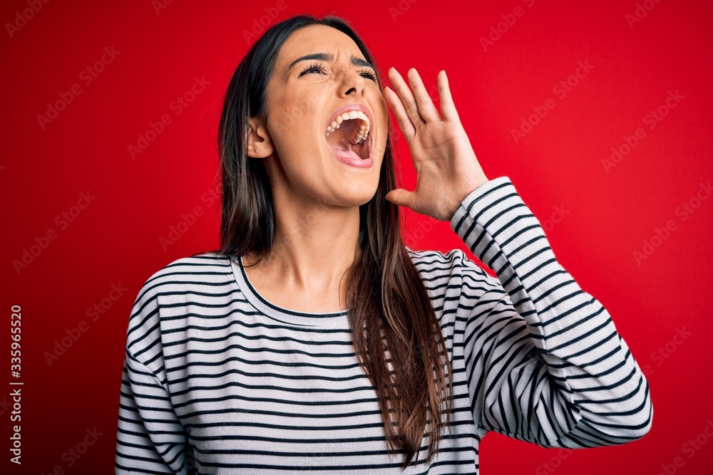 Fototapeta premium Young beautiful brunette woman wearing casual striped t-shirt over red background shouting and screaming loud to side with hand on mouth. Communication concept.