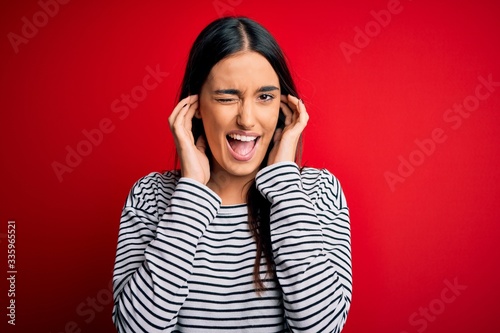 Young beautiful brunette woman wearing casual striped t-shirt over red background covering ears with fingers with annoyed expression for the noise of loud music. Deaf concept. © Krakenimages.com