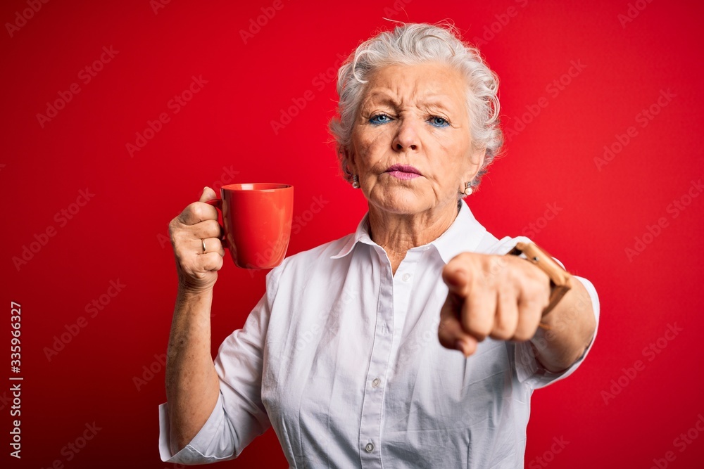 Senior beautiful woman drinking mug of coffee standing over isolated red background pointing with finger to the camera and to you, hand sign, positive and confident gesture from the front Senior beautiful woman drinking mug of coffee standing over isolated red background pointing with finger to the camera and to you, hand sign, positive and confident gesture from the front