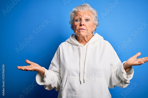 Senior beautiful sporty woman wearing white sweatshirt over isolated blue background clueless and confused expression with arms and hands raised. Doubt concept.