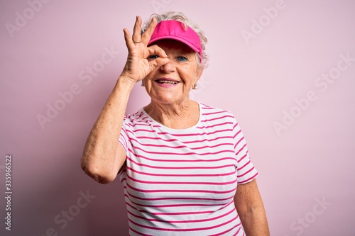 Senior beautiful sporty woman wearing sport cap standing over isolated pink background doing ok gesture with hand smiling, eye looking through fingers with happy face.