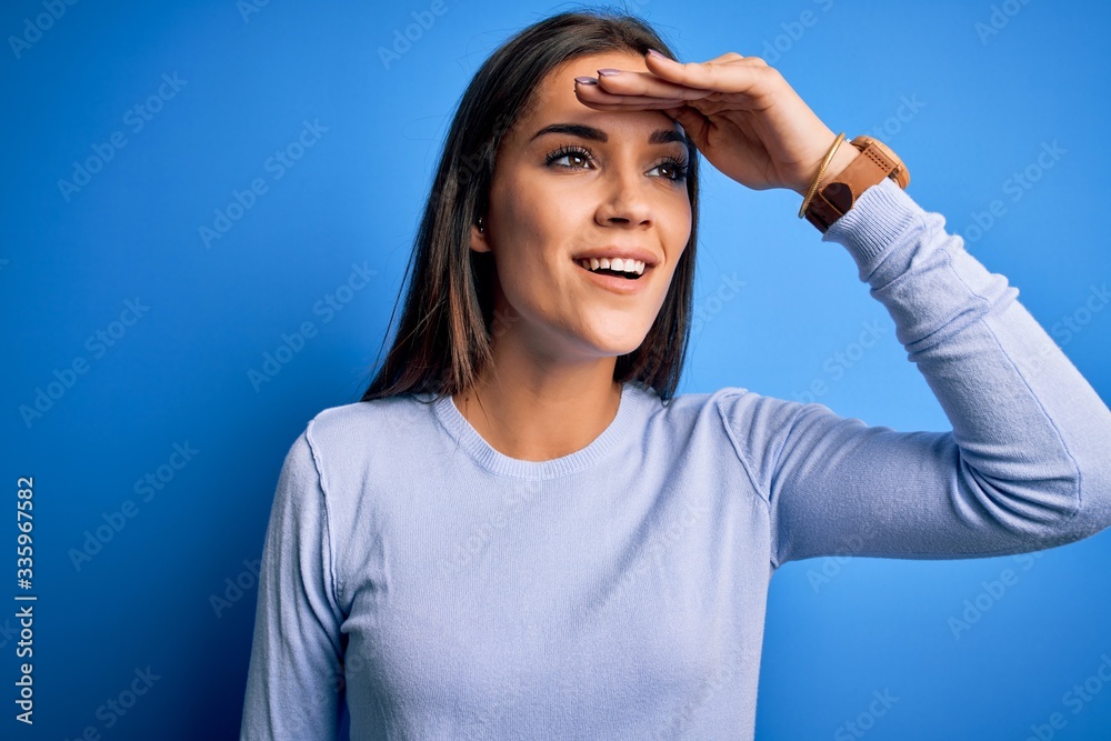 Young beautiful brunette woman wearing casual sweater standing over blue background very happy and smiling looking far away with hand over head. Searching concept.