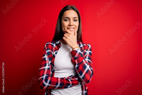 Young beautiful brunette woman wearing casual shirt standing over isolated red background looking confident at the camera smiling with crossed arms and hand raised on chin. Thinking positive.