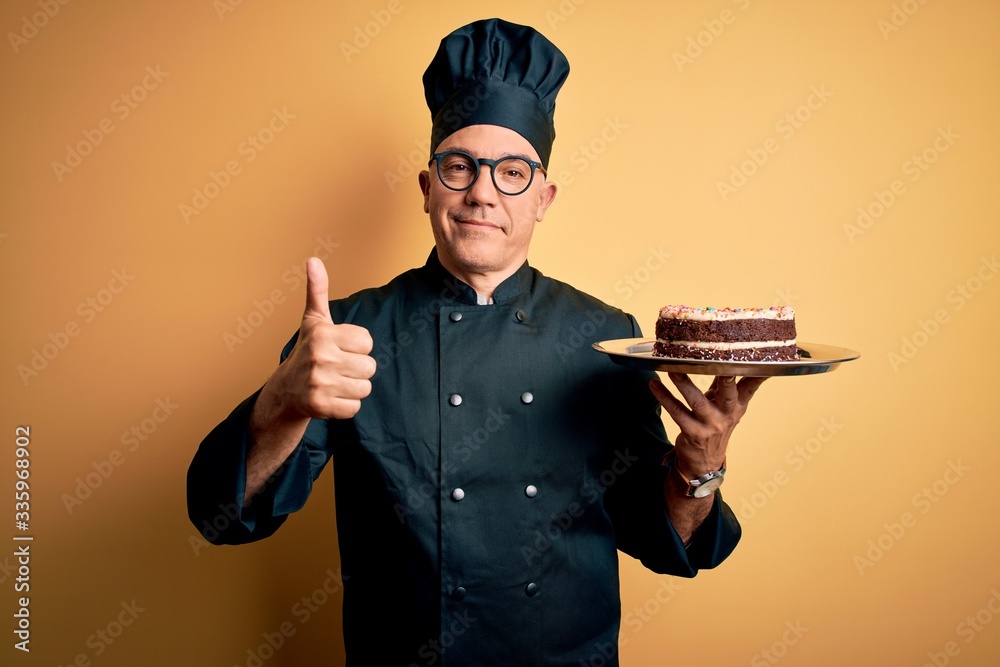 Middle age handsome grey-haired baker man wearing cooker uniform and hat holding cake happy with big smile doing ok sign, thumb up with fingers, excellent sign Middle age handsome grey-haired baker man wearing cooker uniform and hat holding cake happy with big smile doing ok sign, thumb up with fingers, excellent sign