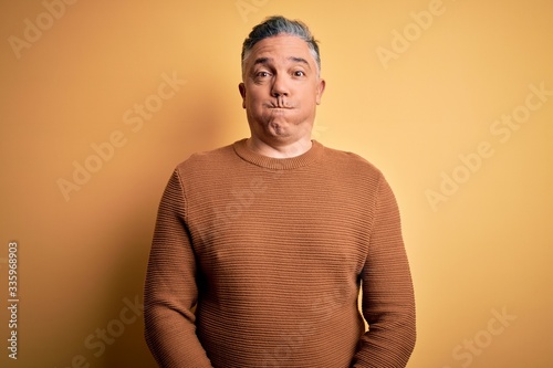 Middle age handsome grey-haired man wearing casual sweater over yellow background puffing cheeks with funny face. Mouth inflated with air, crazy expression.