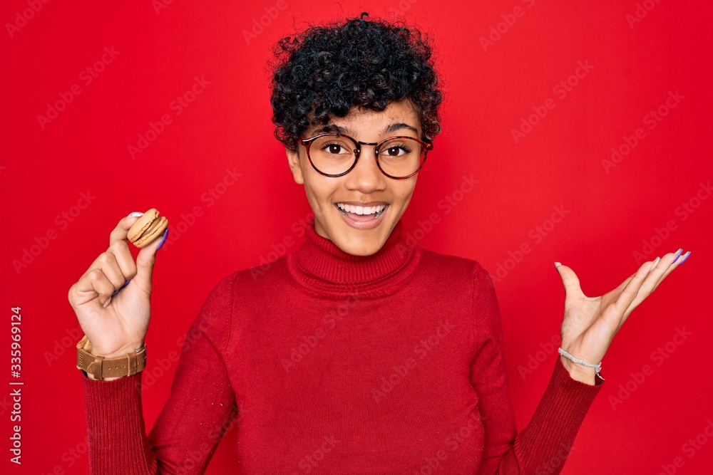 Young beautiful african american afro woman holding chocolate french dessert macaron very happy and excited, winner expression celebrating victory screaming with big smile and raised hands Young beautiful african american afro woman holding chocolate french dessert macaron very happy and excited, winner expression celebrating victory screaming with big smile and raised hands