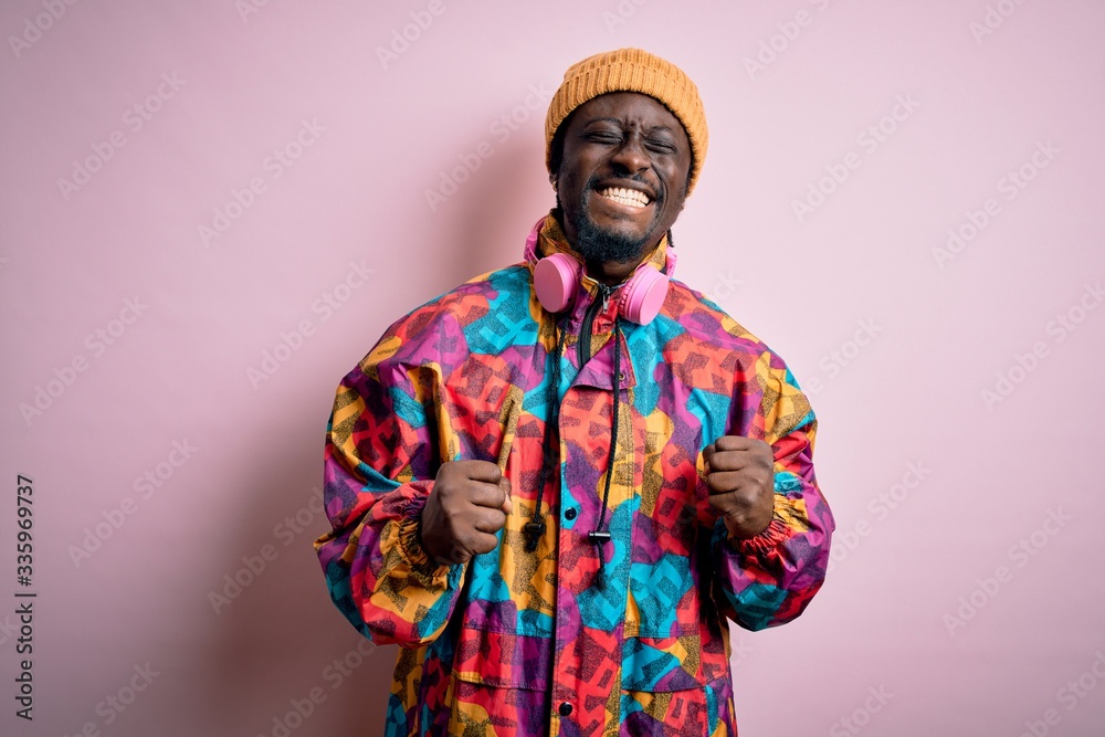 Young handsome african american man wearing colorful coat and cap over pink background excited for success with arms raised and eyes closed celebrating victory smiling. Winner concept. Young handsome african american man wearing colorful coat and cap over pink background excited for success with arms raised and eyes closed celebrating victory smiling. Winner concept.