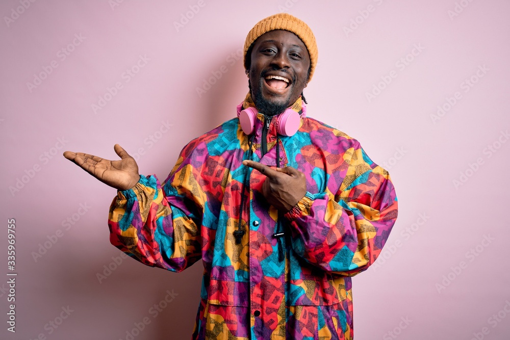 Young handsome african american man wearing colorful coat and cap over pink background amazed and smiling to the camera while presenting with hand and pointing with finger.