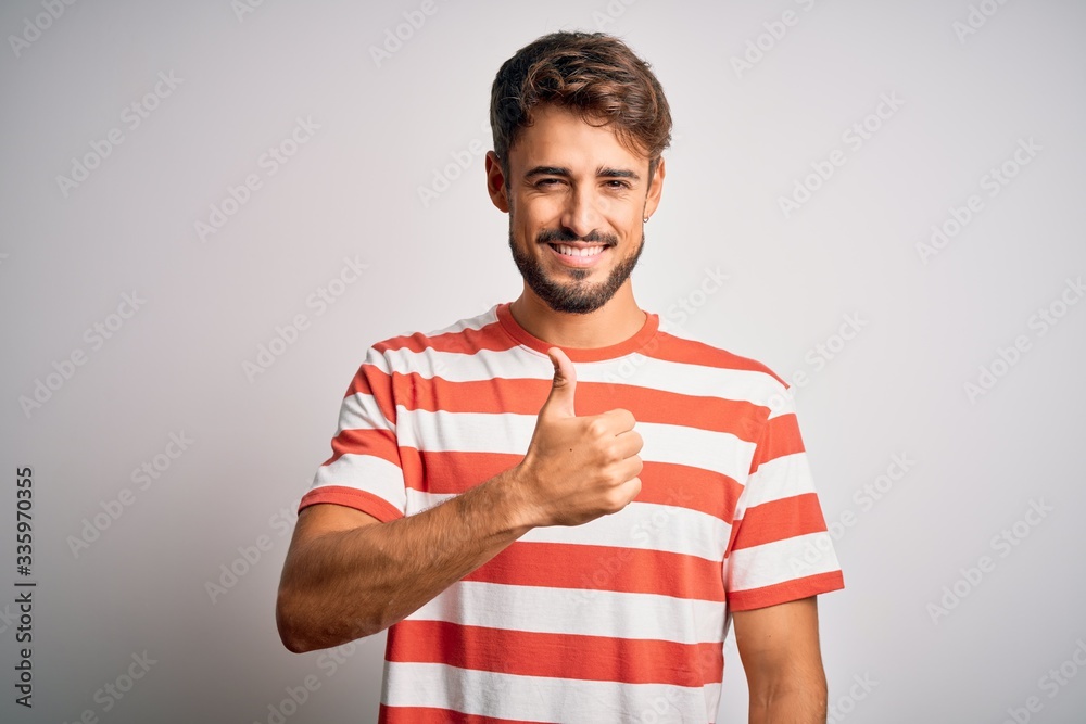 Young handsome man with beard wearing striped t-shirt standing over white background doing happy thumbs up gesture with hand. Approving expression looking at the camera showing success. Young handsome man with beard wearing striped t-shirt standing over white background doing happy thumbs up gesture with hand. Approving expression looking at the camera showing success.