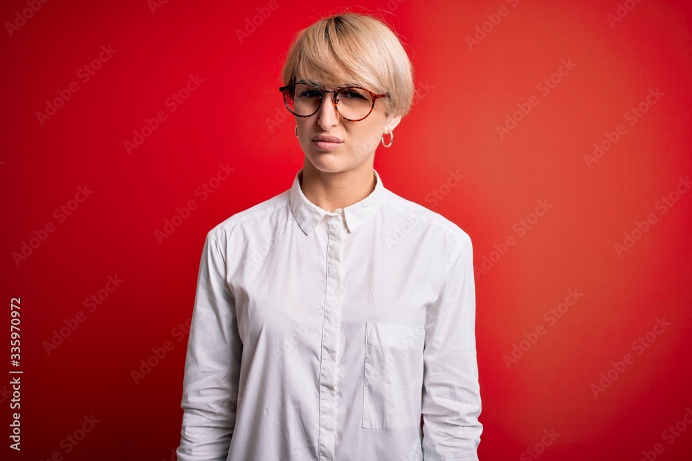 Young blonde business woman with short hair wearing glasses over red background skeptic and nervous, frowning upset because of problem. Negative person.