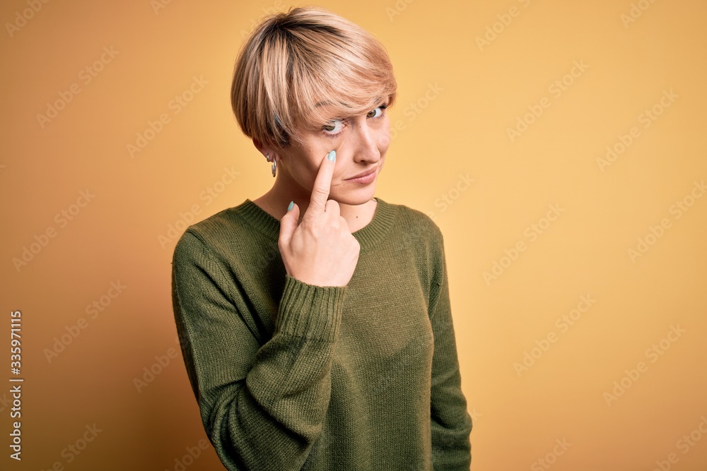 Young blonde woman with modern short hair wearing casual sweater over yellow background Pointing to the eye watching you gesture, suspicious expression Young blonde woman with modern short hair wearing casual sweater over yellow background Pointing to the eye watching you gesture, suspicious expression