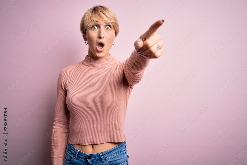 Young blonde woman with short hair wearing casual turtleneck sweater over pink background Pointing with finger surprised ahead, open mouth amazed expression, something on the front