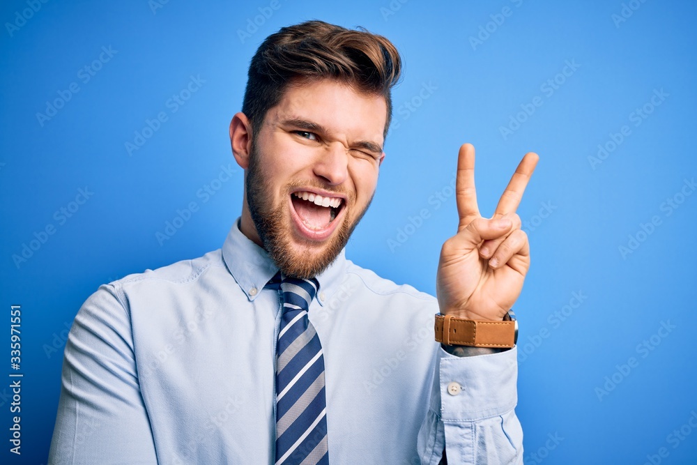 Young blond businessman with beard and blue eyes wearing elegant shirt and tie standing smiling with happy face winking at the camera doing victory sign. Number two.