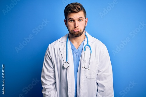 Young blond doctor man with beard and blue eyes wearing white coat and stethoscope depressed and worry for distress, crying angry and afraid. Sad expression.