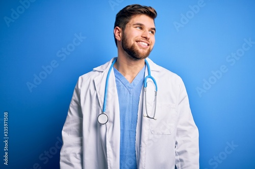 Young blond doctor man with beard and blue eyes wearing white coat and stethoscope looking away to side with smile on face, natural expression. Laughing confident.