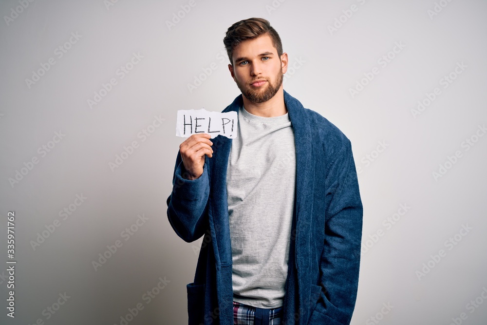 Young blond man with beard and blue eyes wearing pajama holding paper with help message with a confident expression on smart face thinking serious Young blond man with beard and blue eyes wearing pajama holding paper with help message with a confident expression on smart face thinking serious