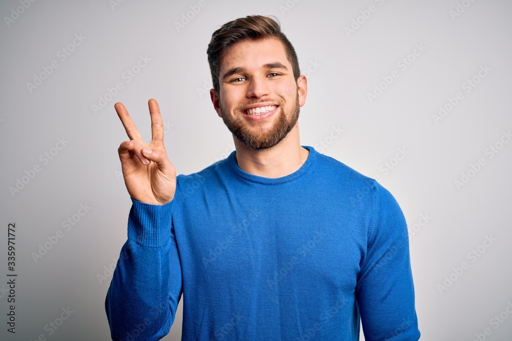Young handsome blond man with beard and blue eyes wearing casual sweater showing and pointing up with fingers number two while smiling confident and happy. Young handsome blond man with beard and blue eyes wearing casual sweater showing and pointing up with fingers number two while smiling confident and happy.